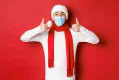 概念一年冠状病毒假期快乐的男人。庆祝一年社会距离穿医疗面具圣诞老人他围巾显示竖起大拇指批准