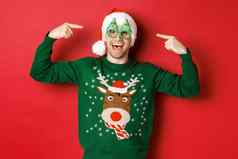 图像兴奋微笑的家伙指出聚会，派对眼镜庆祝一年站快乐的绿色毛衣圣诞老人他红色的背景