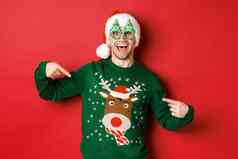 图像快乐男人。聚会，派对眼镜圣诞老人他指出圣诞节毛衣微笑站红色的背景