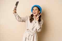 美丽的现代女人摆姿势自拍持有智能手机采取图片照片移动电话站快乐米色背景
