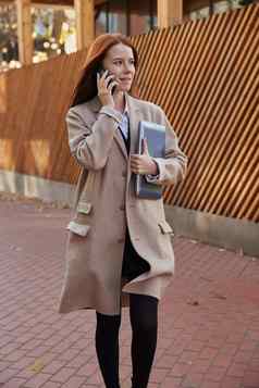 高加索人女人时尚的米色外套调用智能手机持有移动PC