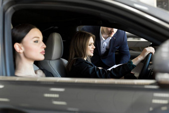 华丽的年轻的女孩坐着内部车微笑相机女孩持有手操舵轮女客户车经销商选择汽车展厅