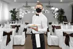 年轻的快乐服务员穿保护脸面具服务食物餐厅