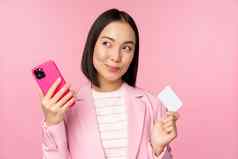 微笑亚洲企业女人夫人西装思考持有智能手机信贷卡计划买smth在线购物移动电话粉红色的背景