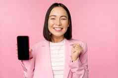 赢得朝鲜文女商人显示智能手机屏幕微笑很高兴展示移动电话应用程序在线商店购物应用程序站粉红色的背景