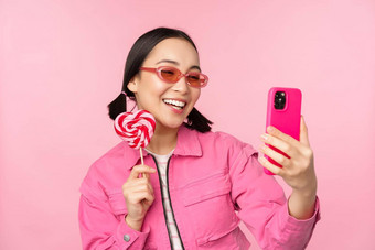 肖像时尚的快乐亚洲女孩采取自拍糖果<strong>萝莉</strong>波普糖果微笑采取照片移动应用程序站粉红色的背景