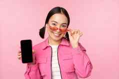 肖像时尚的现代亚洲女孩显示移动电话屏幕智能手机应用程序接口站太阳镜粉红色的背景