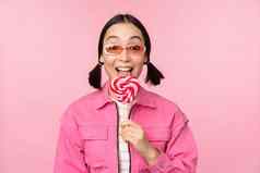 时尚的朝鲜文女孩舔萝莉波普吃糖果微笑站太阳镜粉红色的背景