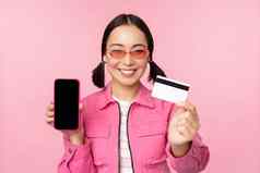图像微笑朝鲜文女人显示信贷卡移动电话屏幕智能手机应用程序接口支付在线购物非接触式站粉红色的背景