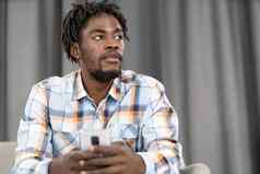 深思熟虑的非洲美国男人。智能手机检查社会媒体更新信息肖像年轻的男人。持有智能手机坐着沙发社会媒体概念