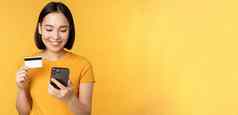 在线购物微笑亚洲女孩信贷卡移动电话应用程序支付非接触式订单智能手机应用程序站黄色的背景