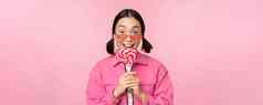愚蠢的可爱的亚洲女模型舔萝莉波普吃糖果甜蜜的微笑兴奋站粉红色的背景