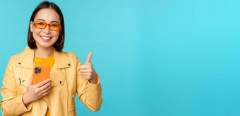 微笑亚洲女人显示拇指记录移动电话智能手机应用程序推荐站蓝色的背景
