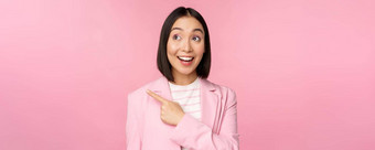 热情的年轻的亚洲女商人办公室员工指出手指左横幅广告快乐微笑显示广告粉红色的背景