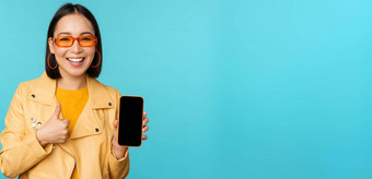 年轻的时尚的中国人女人显示移动电话屏幕拇指智能手机应用程序站蓝色的背景