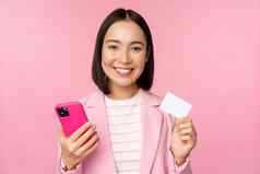 图像微笑快乐亚洲女商人显示信贷卡支付在线智能手机应用程序订单移动方站粉红色的背景