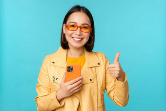 微笑亚洲女人显示拇指记录移动电话智能手机应用程序推荐站蓝色的背景