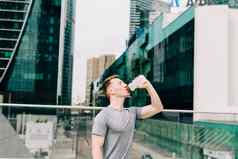 累了年轻的男人。跑步者喝水放松体育运动培训持有水瓶健身锻炼夏天城市城市街多云的天空