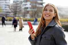 肖像微笑女人穿灰色的夹克持有智能手机行人街