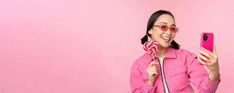肖像时尚的快乐亚洲女孩采取自拍糖果<strong>萝莉</strong>波普糖果微笑采取照片移动应用程序站粉红色的背景