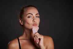 有吸引力的中间岁的欧洲女人健康的新鲜的光滑的晒黑皮肤摆姿势刮痧沙玉石头按摩器面部提升淋巴排水按摩孤立的黑色的背景