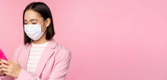 亚洲女商人医疗脸面具移动电话日本语女售货员企业夫人西装持有智能手机站粉红色的背景