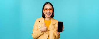 年轻的时尚的中国人女人显示移动电话屏幕拇指智能手机应用程序站蓝色的背景
