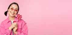 微笑亚洲女人太阳镜持有萝莉波普糖果吃糖果快乐站粉红色的背景