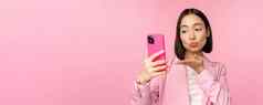 时尚的亚洲女商人女孩西装采取自拍智能手机视频闲谈，聊天移动电话应用程序摆姿势粉红色的工作室背景
