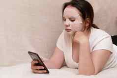 年轻的女人面具化妆品织物谎言沙发白色t恤冲浪智能手机概念检疫首页护理