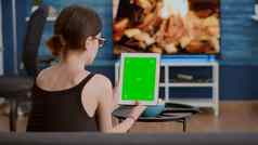 年轻的女人坐着沙发持有垂直数字平板电脑绿色屏幕看在线社会媒体内容