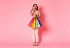 完整的长度肖像逗乐快乐快乐的亚洲女孩触碰脸颊微笑持有购物袋购买很多的事情折扣销售站粉红色的背景