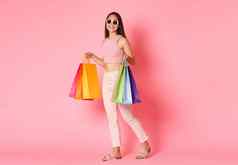 完整的长度肖像华丽的亚洲女购物携带袋夏天衣服走商店折扣穿太阳镜站粉红色的背景