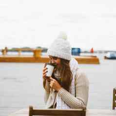 美丽的年轻的女孩喝咖啡茶塑料杯子秋天冬天女人长头发温暖的衣服坐着海滨波罗的海海港口温暖热喝