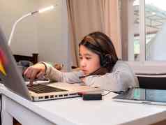 亚洲女孩耳机坐桌子上研究在线移动PC孩子穿耳机笔迹笔记本学习互联网教训检疫女孩学生学习虚拟互联网在线类学校由于科维德流感大流行