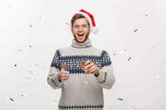 圣诞概念快乐年轻的高加索人胡子男人。持有香槟玻璃五彩纸屑背景庆祝圣诞节一天