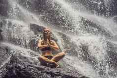 健康水疗中心假期瑜伽冥想概念年轻的女人坐着莲花位置岩石热带瀑布