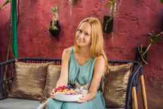 年轻的女人地中海早餐坐着沙发猫吃健康的热带早餐奶昔碗热带水果装饰模式色彩斑斓的酸奶姜黄螺旋藻装饰水果花传讲种子椰子格兰诺拉麦片菠萝薄荷草莓