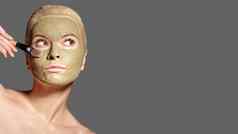 美丽的女人应用绿色面部面具美治疗水疗中心女孩应用粘土面部面具灰色背景