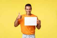 快乐英俊的拉美裔的家伙橙色t恤持有空白纸一块微笑批准显示翘拇指手势促销推荐访问公司网络页面黄色的背景