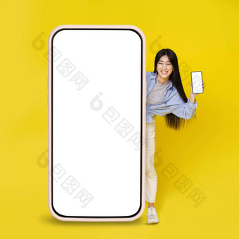 可爱的亚洲年轻的女孩兴奋窥视大巨大的垂直细胞电话持有小手白色空白屏幕孤立的黄色的背景伟大的提供产品放置复制空间