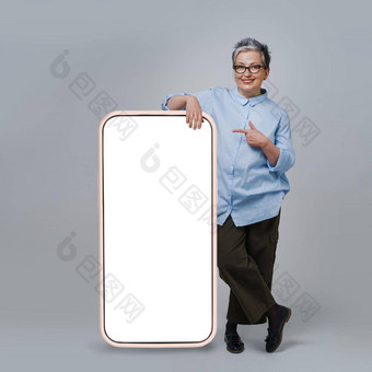 成熟的灰色头发女人指出手指屏幕巨大的巨大的智能手机白色空白屏幕站靠穿休闲孤立的白色背景免费的空间模拟