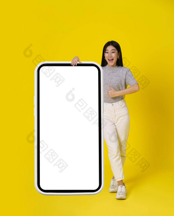 迷人的亚洲女孩手势拇指站巨大的巨大的智能手机白色屏幕快乐穿着休闲孤立的黄色的背景免费的空间模拟产品放置移动应用程序添加