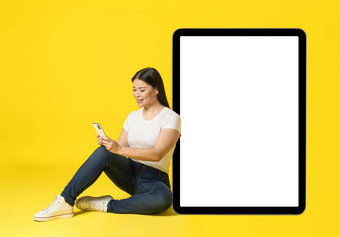 亚洲女人靠<strong>巨</strong>大的<strong>巨</strong>大的平板电脑坐着地板上电话手休闲白色屏<strong>幕</strong>电话移动应用程序广告孤立的黄色的产品放置