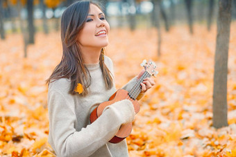 年轻的美丽的混合比赛女人玩尤克里里琴唱歌秋天公园