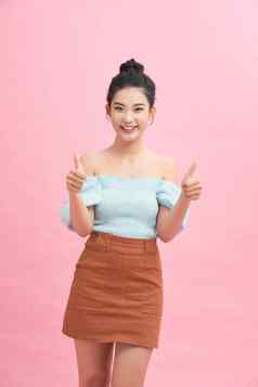 漂亮的亚洲女人站粉红色的背景微笑信号拇指