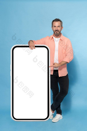 中间岁的灰色头发的男人。靠巨大的巨大的智能手机指出白色屏幕快乐微笑相机穿桃子衬衫孤立的蓝色的背景成熟的适合男人。智能手机