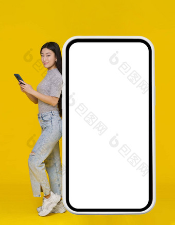 微笑亚洲女孩电话手靠回来巨大的巨大的智能手机数字平板电脑白色黑色的屏幕站穿休闲孤立的黄色的背景免费的空间模拟