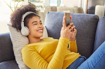 年轻的快乐的混合比赛女人穿耳机听音乐打字消息电话首页快乐拉美裔女卷曲的非洲式发型社会媒体手机放松沙发上首页