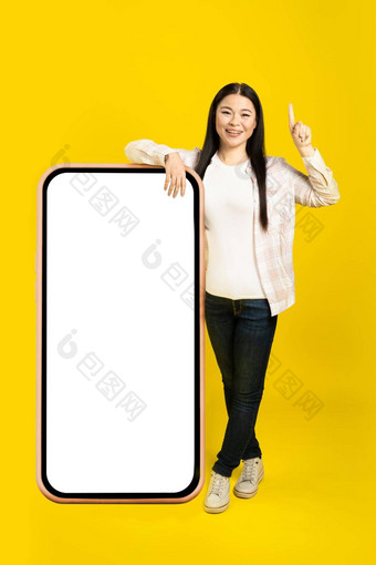 指出手指中间年龄亚洲女人靠巨大的巨大的智能手机空白白色屏幕穿休闲装孤立的黄色的背景免费的空间模拟
